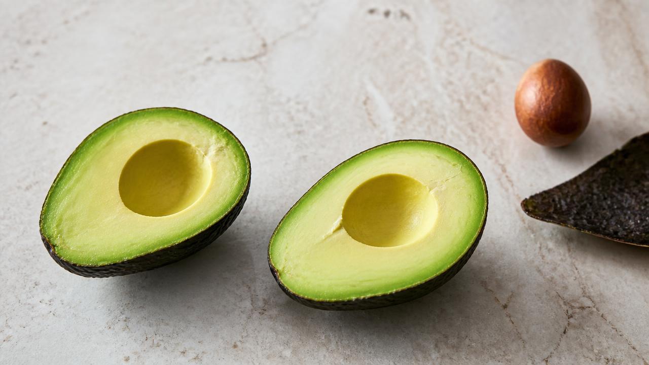 Two halves of a ripe avocado with the flesh exposed beside the separated brown pit and discarded skin on a pale stone surface