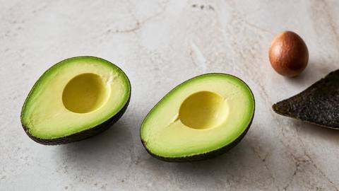 Two halves of a ripe avocado with the flesh exposed beside the separated brown pit and discarded skin on a pale stone surface