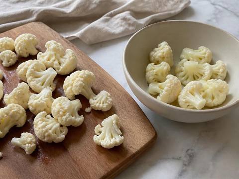 Small plain raw cauliflower florets on a wooden board beside a bowl of plainly steamed florets in a home kitchen