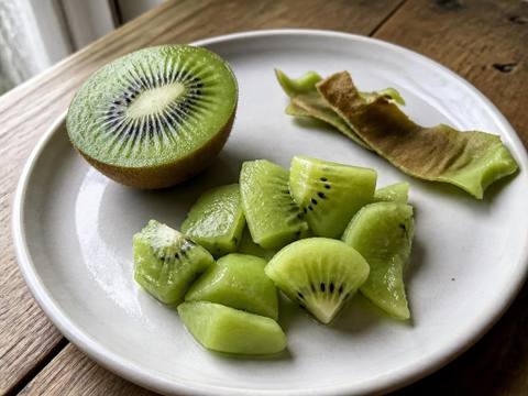 A ripe kiwi cut in half beside irregular peeled kiwi pieces and a strip of discarded brown peel on a pale ceramic plate