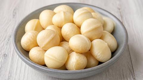 A grey ceramic bowl of plain shelled macadamia nuts on a pale wooden table
