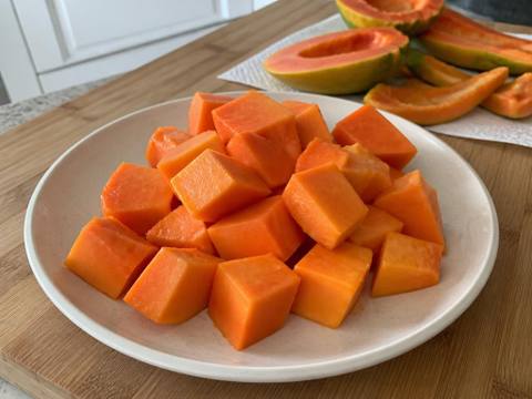 Cubes of ripe orange papaya flesh on a pale ceramic plate with the skin and scooped-out seeds set aside separately on a wooden board