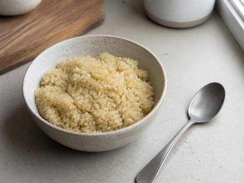 A small bowl of plain fully cooked fluffy quinoa on a kitchen counter with a spoon resting beside it