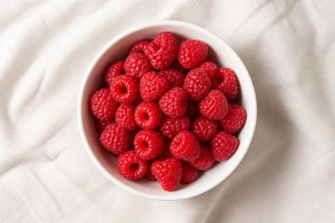 Fresh red raspberries in a small white bowl on a cream linen surface