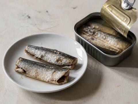 Two plain sardines packed in water on a small white plate beside an open plain tin of sardines in water